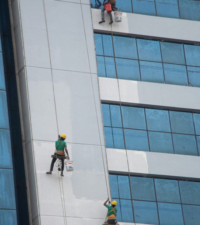 Tres trabajadores que limpian cristales colgados con arneses desde un edificio muy alto.